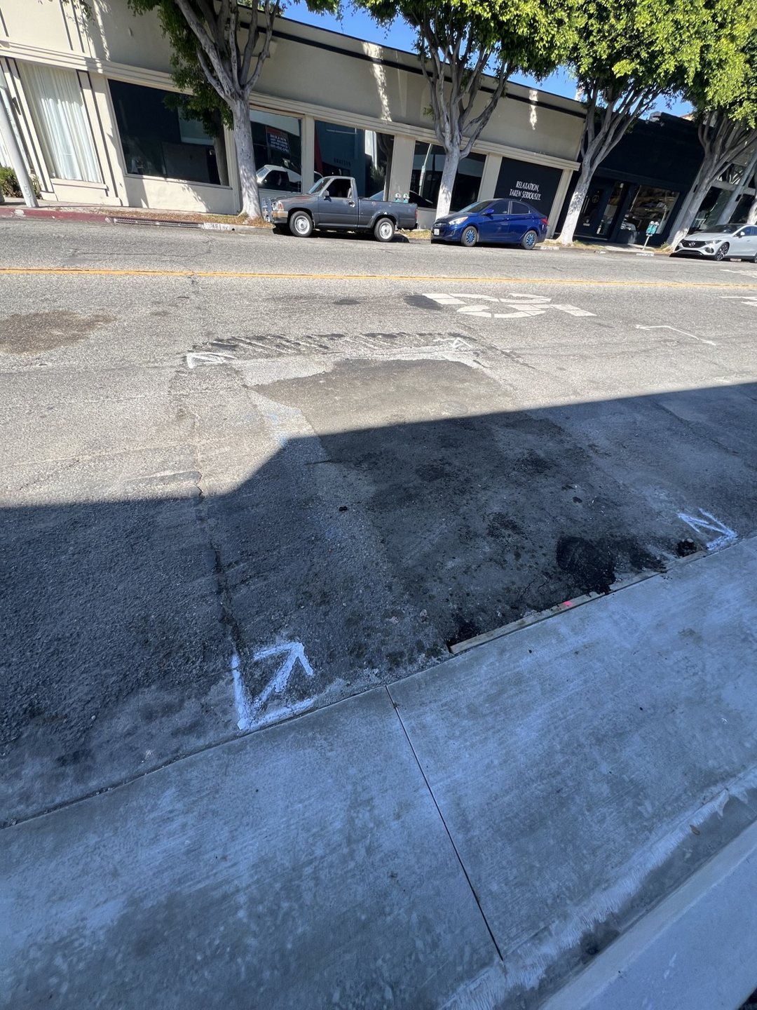 Asphalt patch and utility markings in the roadway beside the project sidewalk.