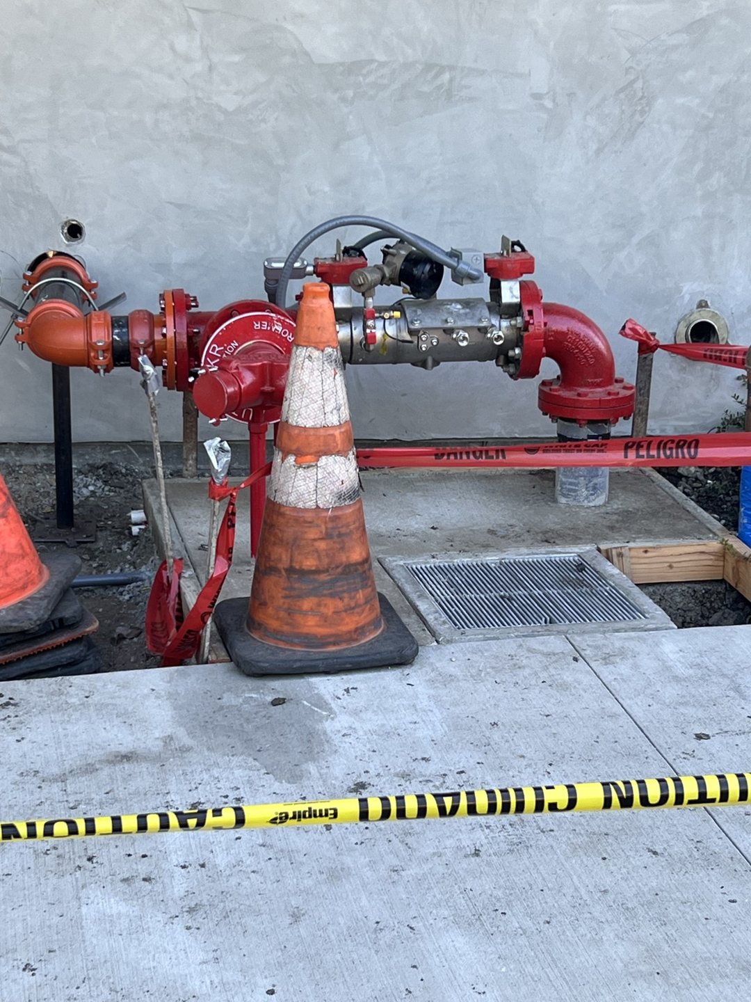 Red piping and valve assembly beside the building, with cones and caution tape marking the work area.